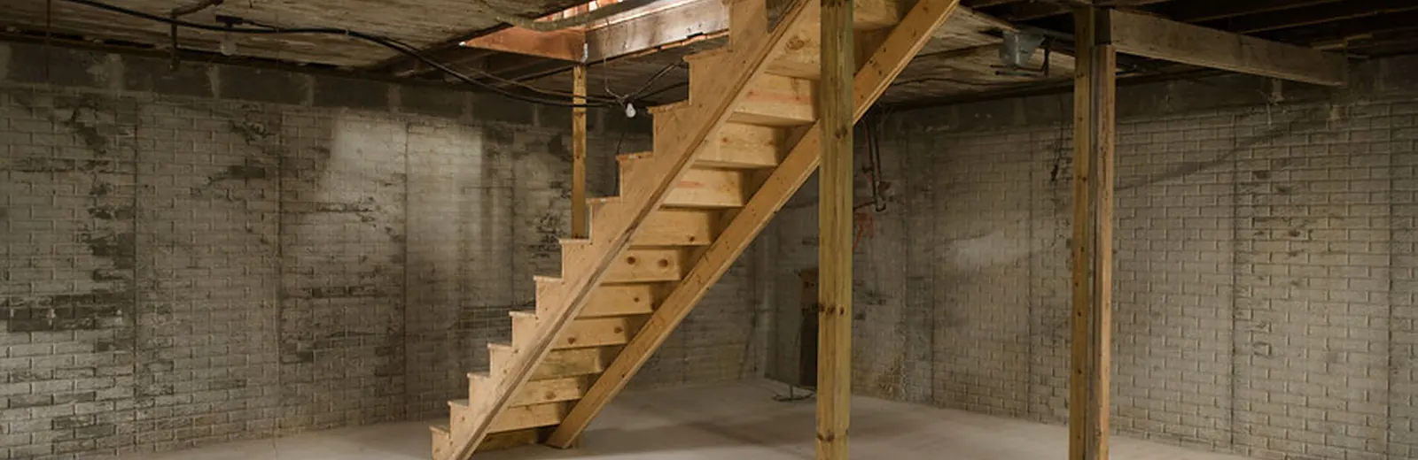 Unfinished basement with exposed foundation walls and wooden stairs